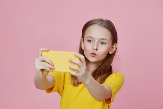 Светловолосая девочка в жёлтой футболке делает селфи на розовом фоне