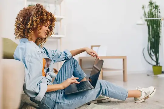 Кудрявая девушка в джинсовой куртке работает за ноутбуком на полу