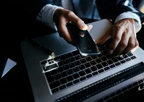 Мужчина в рубашке держит смартфон над клавиатурой ноутбука