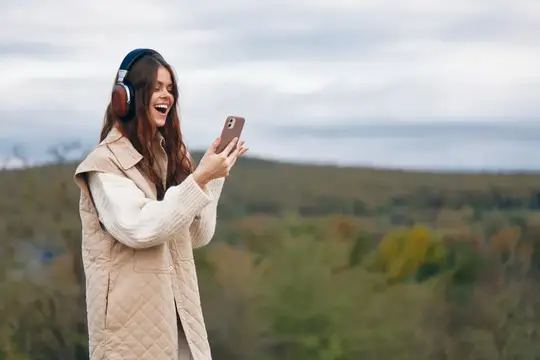 Смеющаяся девушка в наушниках смотрит в телефон на природе