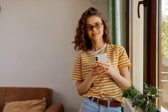 Улыбающаяся девушка в очках с телефоном у окна