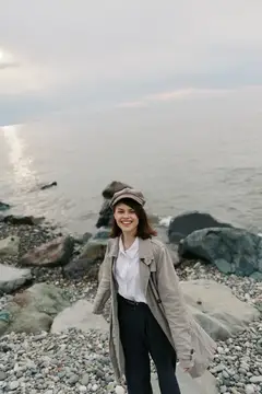 Улыбающаяся девушка в кепке и плаще стоит на каменистом берегу моря