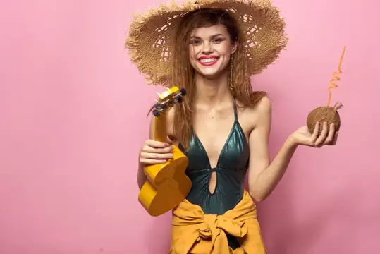 Улыбающаяся девушка в соломенной шляпе с укулеле и коктейлем на розовом фоне