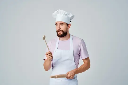 Бородатый мужчина в поварском колпаке и фартуке держит деревянную ложку и скалку