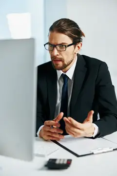 Сосредоточенный мужчина в очках и костюме жестикулирует перед монитором в офисе