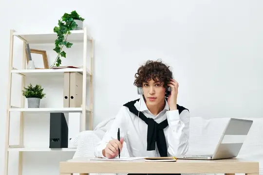 Кудрявая девушка в белой рубашке с наушниками работает за ноутбуком дома