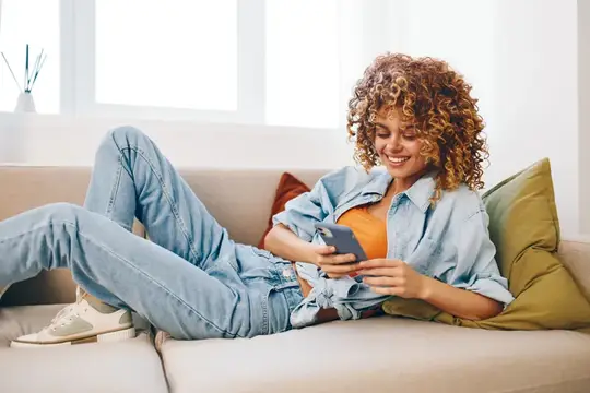 Кудрявая девушка лежит на диване со смартфоном и улыбается