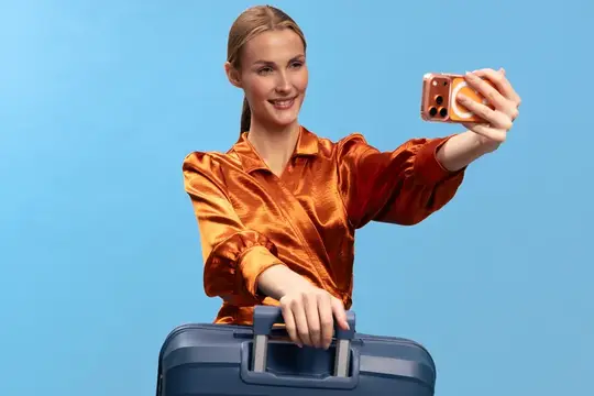 Улыбающаяся женщина с чемоданом в оранжевой рубашке делает селфи на голубом фоне