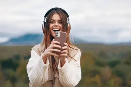 Улыбающаяся девушка в наушниках смотрит в телефон на природе