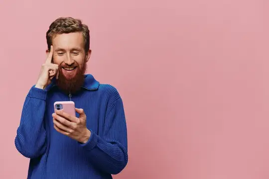 Бородатый мужчина в синем свитере с телефоном на розовом фоне