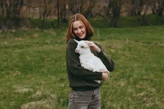 Женщина улыбается, поддерживая козленка на открытом воздухе, обстановка на открытом воздухе для повествования об образе жизни, привязанность, объятия и природа, с мягким светом, легким ветерком и…