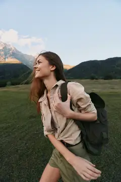 Жизнерадостная женщина, ведущая активный образ жизни на природе, исследует тропу в легкой рубашке и шортах, с рюкзаком, волосы развеваются на ветру, обширный пейзаж, яркая зелень, ясное небо…