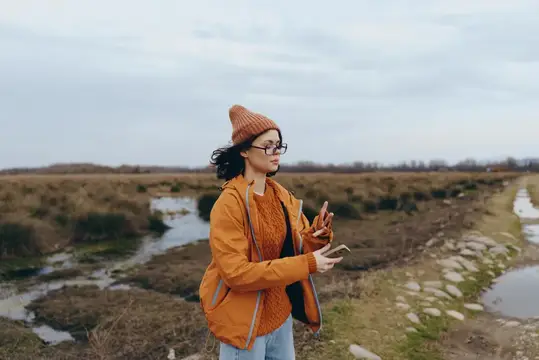 Молодая женщина в оранжевой куртке и шапке смотрит в телефон на фоне осеннего поля