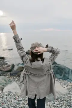 Вид со спины на женщину в клетчатом пальто и шляпе, поднимающую руки на каменистом берегу у спокойного моря под пасмурным небом