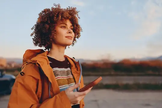 Кудрявая девушка в оранжевой куртке смотрит вдаль с телефоном в руке