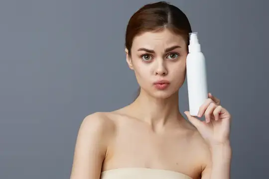 Молодая женщина с белым флаконом косметики у лица на сером фоне