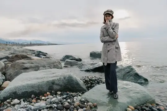 Молодая женщина в плаще и кепке стоит на камнях у моря