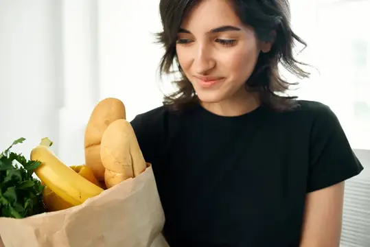 Молодая женщина с бумажным пакетом продуктов улыбается дома