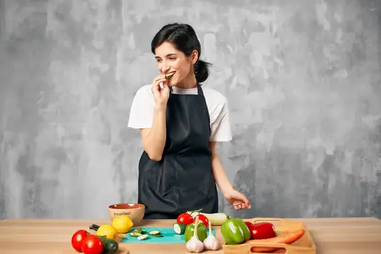 Улыбающаяся женщина в фартуке пробует овощ на кухне
