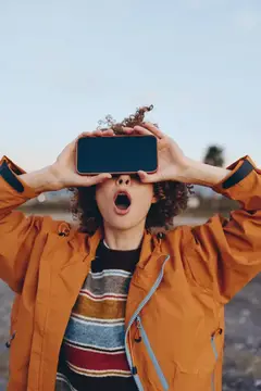 Кудрявая девушка в оранжевой куртке с телефоном у лица удивлённо открыла рот