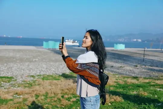Улыбающаяся азиатская девушка фотографирует море на смартфон