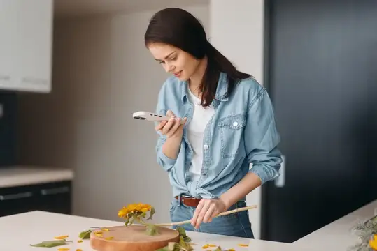 Молодая женщина фотографирует цветок на телефон в светлой кухне