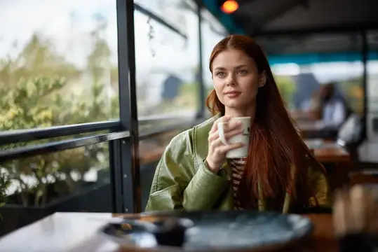 Рыжеволосая девушка с чашкой кофе у окна кафе