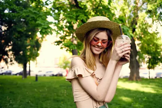 Улыбающаяся блондинка в соломенной шляпе с напитком в парке