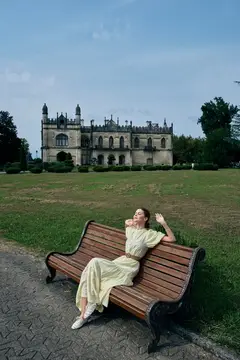 Молодая женщина в светлом платье отдыхает на скамейке перед старинным замком