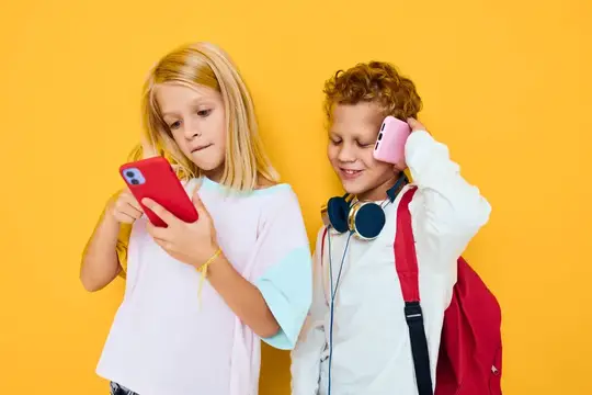 Белокурая девочка и кудрявый мальчик со смартфонами на жёлтом фоне