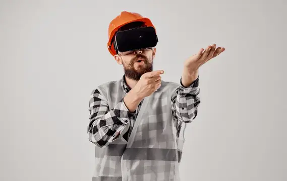 Бородатый мужчина в каске и VR-очках жестикулирует на сером фоне
