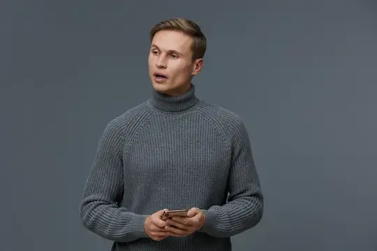 Удивлённый парень в серой водолазке с телефоном на сером фоне