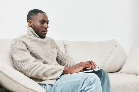 Темнокожий мужчина в свитере работает за ноутбуком на диване