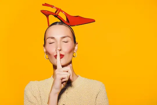 Модная женщина с красной туфлей на голове делает жест молчания, прижав палец к губам