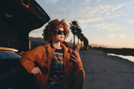 Женщина на улице на закате, отдыхает у машины, куртка и кудрявые волосы, телефон в руке, расслаблена на дороге, небо золотого часа, теплые цвета, боке