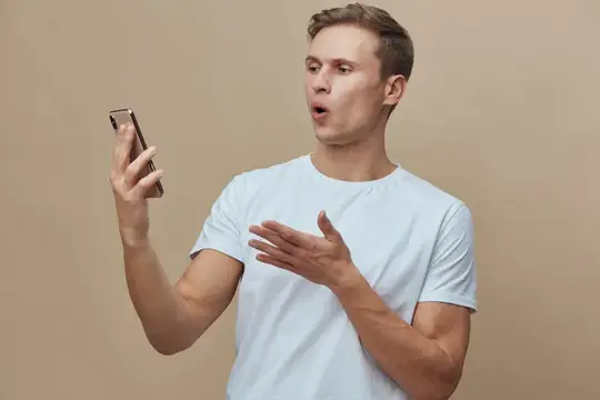Удивлённый парень в белой футболке смотрит на телефон на бежевом фоне