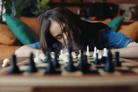 Молодая женщина играет в шахты, и изо рта у неё идёт дым; она сидит перед деревянным столом и диваном