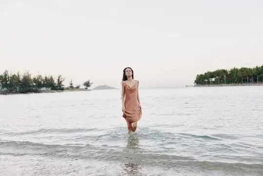 Молодая женщина в бежевом платье стоит в воде на фоне моря и берега