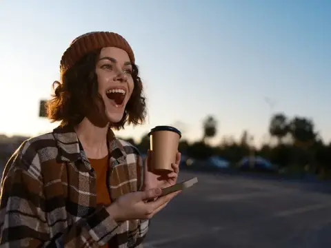 Смеющаяся девушка в шапке с кофе и телефоном на закате