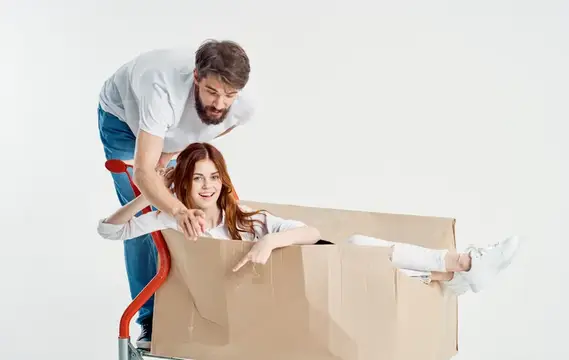 Мужчина везёт улыбающуюся рыжеволосую девушку в картонной коробке на тележке
