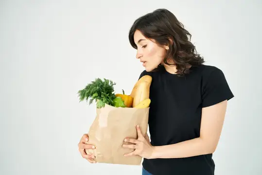 Молодая женщина с бумажным пакетом продуктов смотрит на свежие овощи