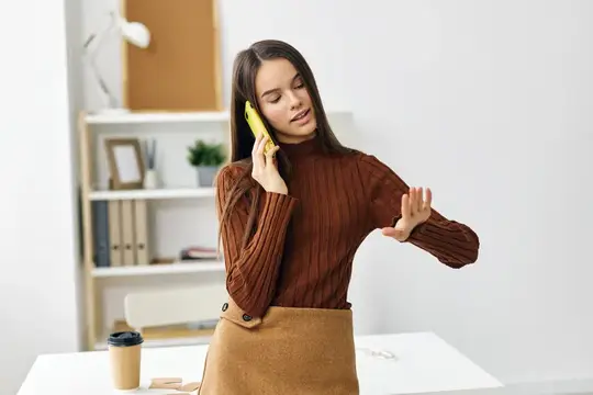 Молодая женщина в коричневом свитере разговаривает по жёлтому телефону в офисе