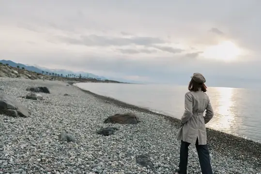Женщина в плаще стоит на галечном берегу и смотрит на море