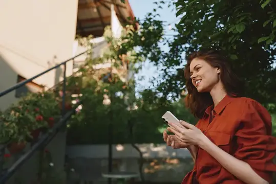 Уличная лайфстайл-сцена с женщиной: естественная улыбка, повседневная мода, телефон в руке, ярко-красная рубашка, залитая солнцем садовая атмосфера, расслабленно и радостно