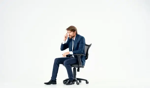 Уставший бородатый мужчина в деловом костюме сидит на офисном стуле и трет переносицу