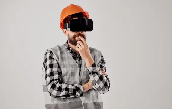 Бородатый мужчина в оранжевой каске и VR-очках задумчиво смотрит