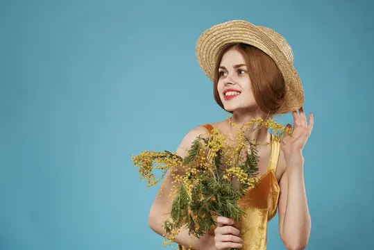 Улыбчивая рыжеволосая девушка в соломенной шляпе с букетом мимозы на голубом фоне