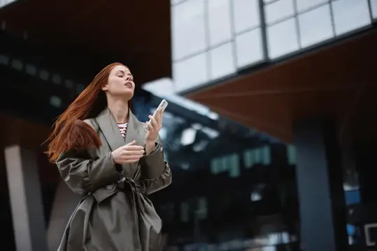Рыжеволосая девушка в плаще со смартфоном у офисного здания