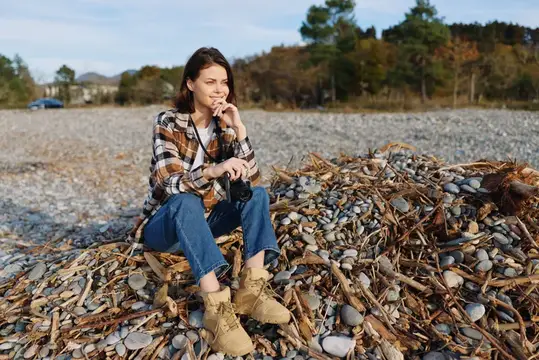 Молодая женщина с фотокамерой сидит на камнях у моря