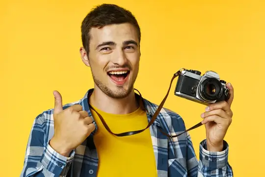 Улыбающийся парень с камерой показывает большой палец вверх на жёлтом фоне